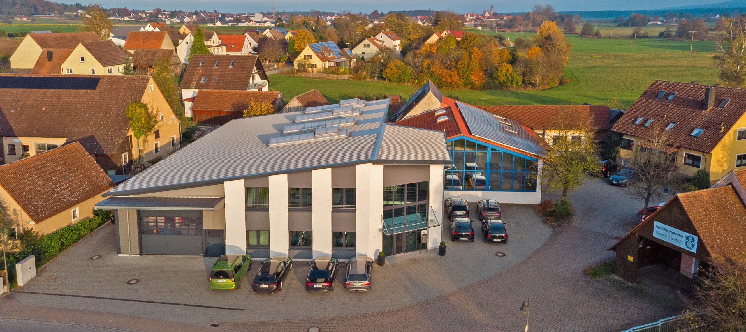 Blick auf das Firmengebäude der WU Mechanische Werkstätte GmbH mit mehreren geparkten Autos davor, umgeben von Wohnhäusern und grüner Landschaft.