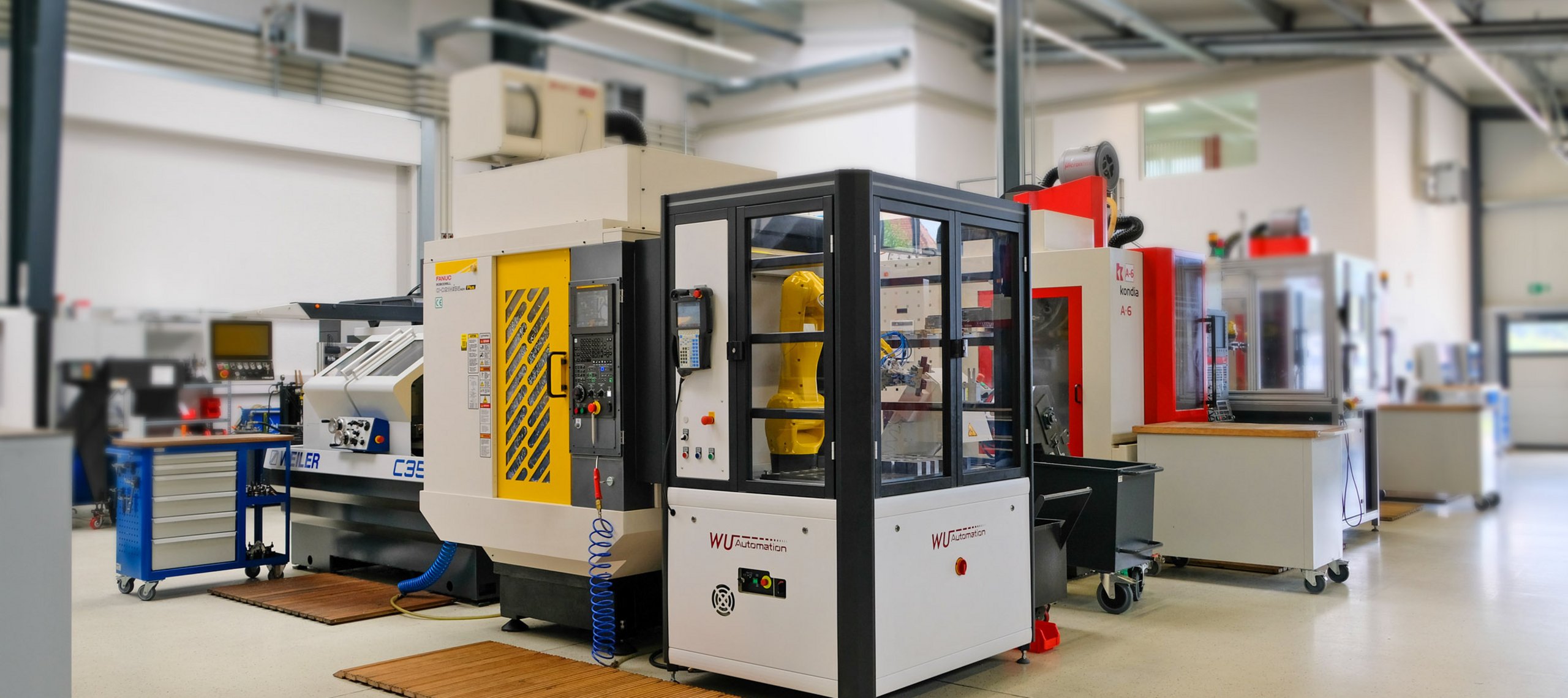 Fanuc Robodrill - Automatisiertung - WU Systems Industrielle Fertigungsanlage 'Fanuc Robodrill' mit gelbem Roboterarm in geschütztem Gehäuse, beschriftet mit 'WU-SYSTEMS' in einer Werkhalle der WU Mechanische Werkstätte GmbH.