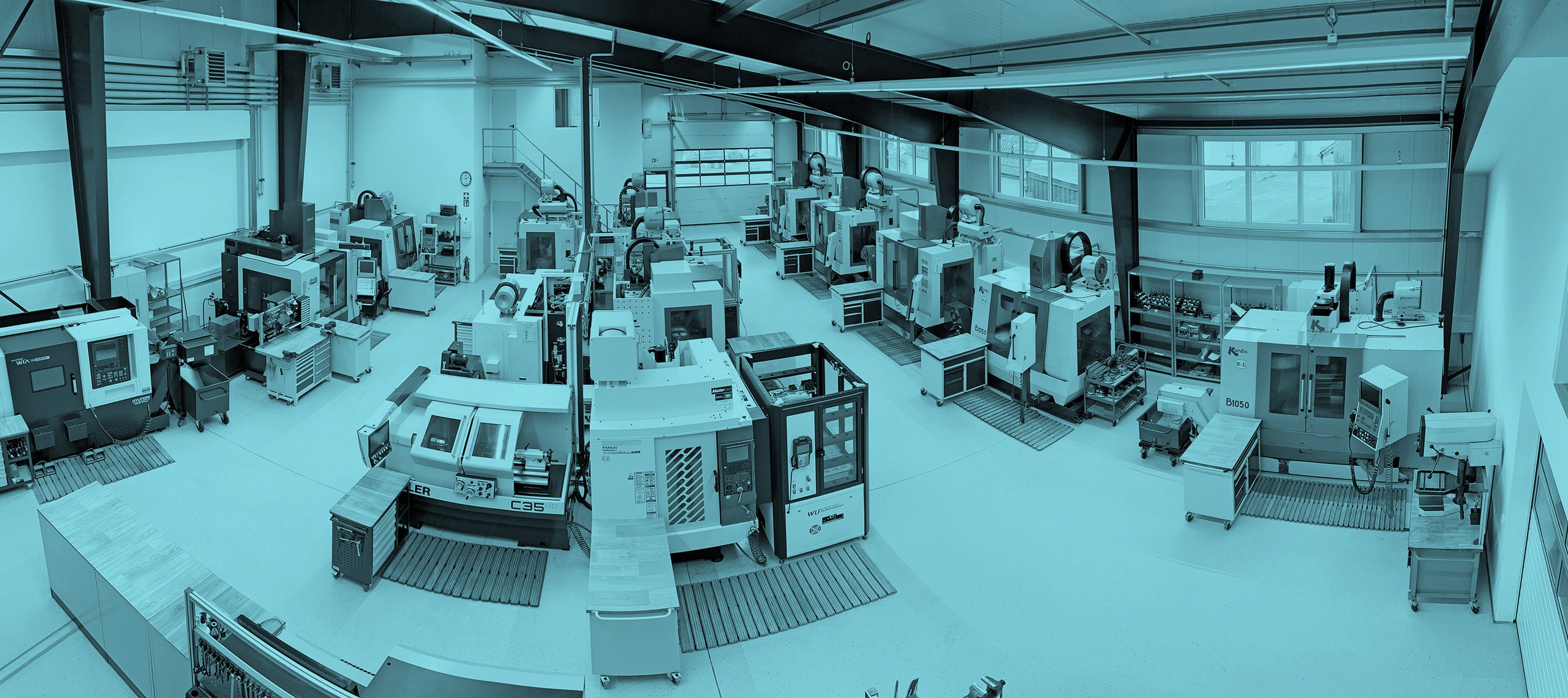 Blick in eine Werkhalle mit mehreren CNC-Fräsmaschinen in Reihen, Arbeitsplätze mit blauen Schubladenschränken, helle Wände und große Fenster.
