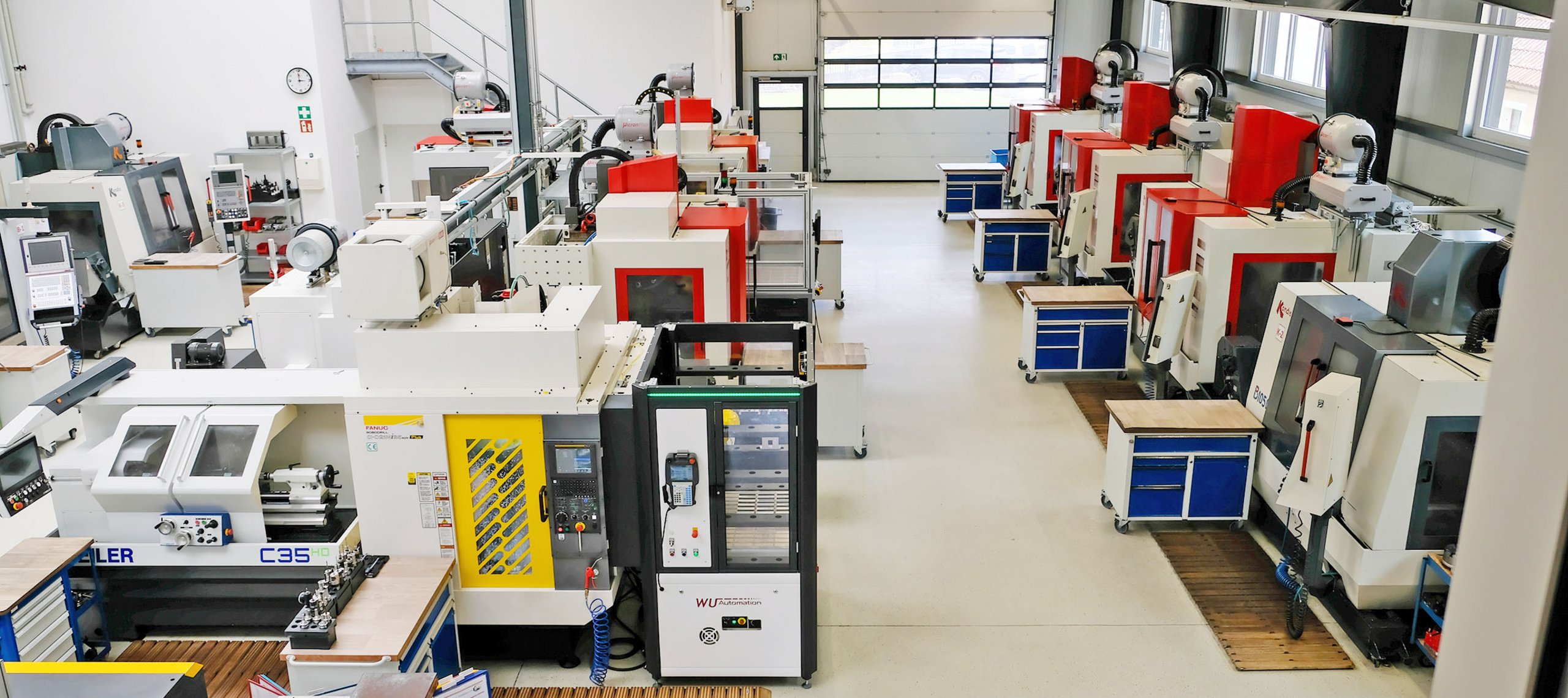 Blick in eine Werkhalle mit mehreren CNC-Fräsmaschinen in Reihen, Arbeitsplätze mit blauen Schubladenschränken, helle Wände und große Fenster.